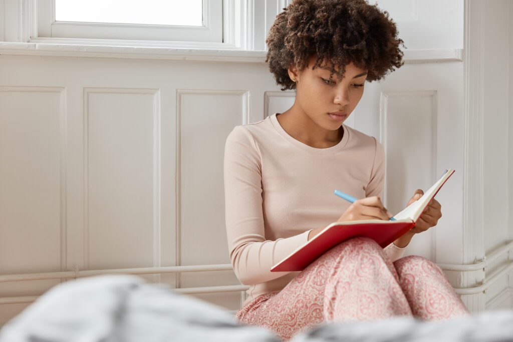 Young woman journaling during an emotional self-check for woman, sitting quietly by a window and reflecting in a notebook.